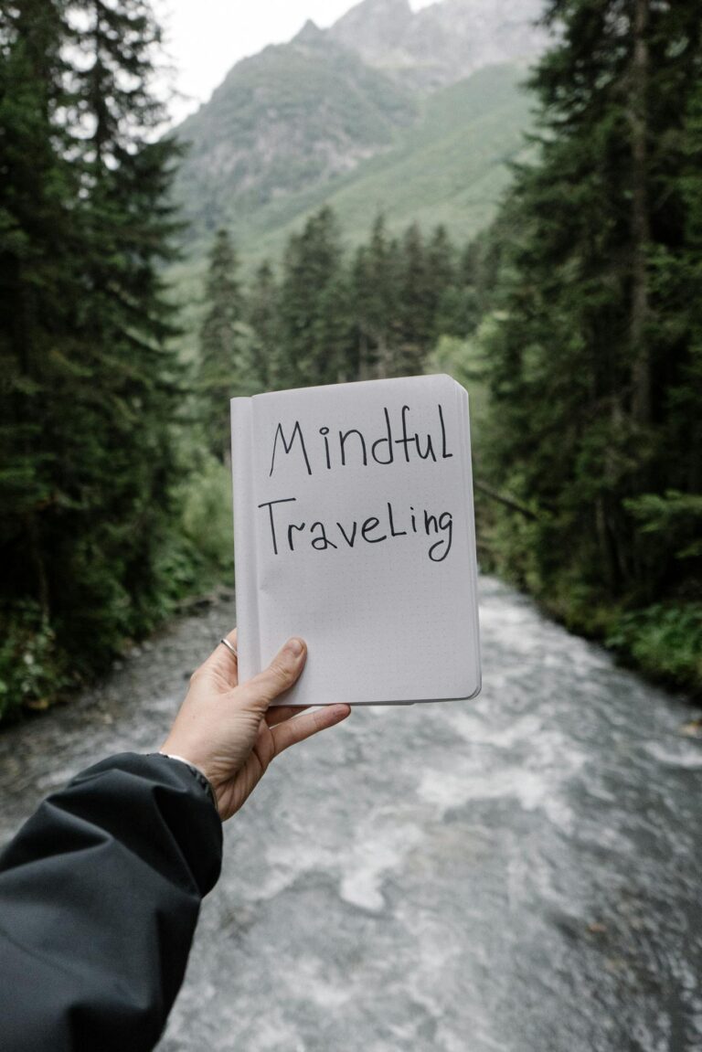 A person holds a notebook titled 'Mindful Traveling' against a backdrop of a serene forest and river.