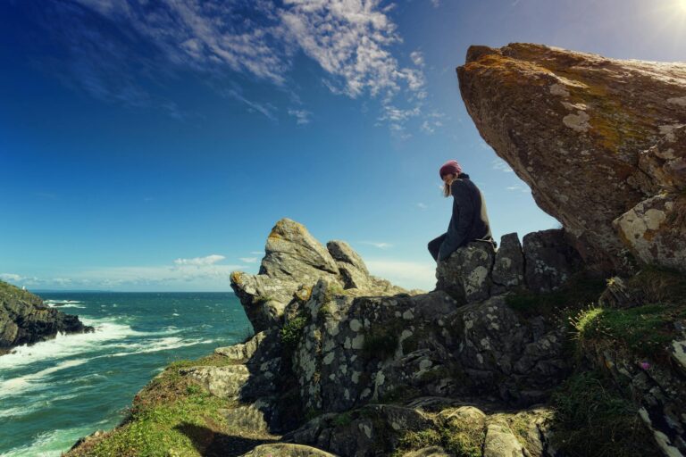 A person sits on a rocky cliff by the seaside, contemplating the vast ocean under a bright blue sky.