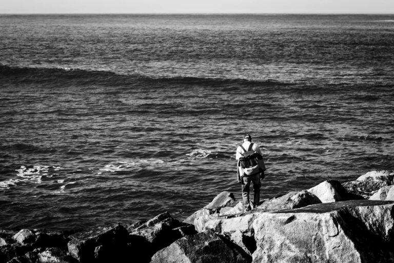 Black and white photo of a person standing on rocks by the ocean. Tranquil and introspective.