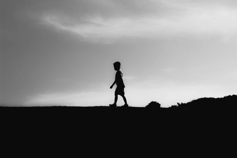 Silhouette of a young boy walking along a hillside, captured in black and white against a dramatic sky.